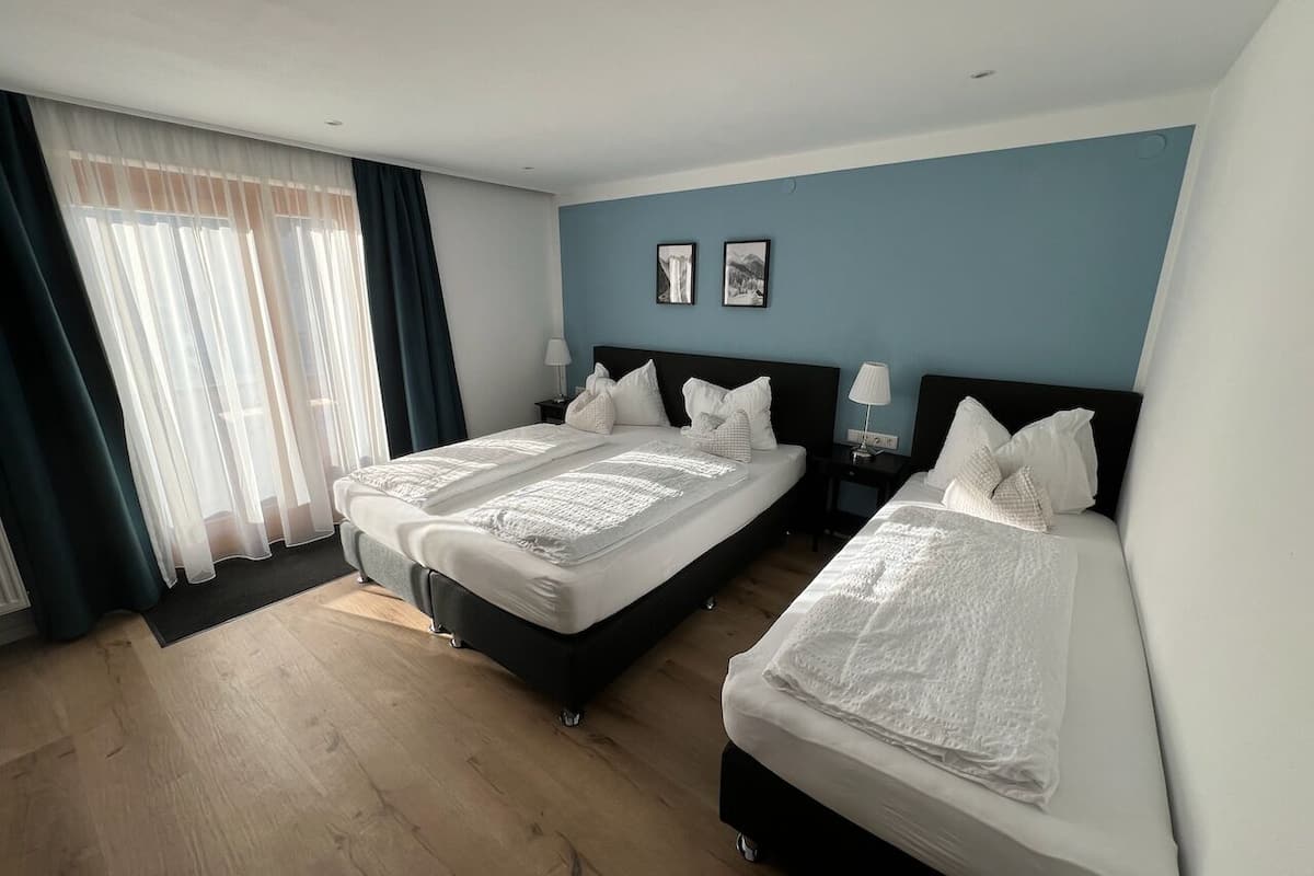 Une chambre d’hôtel avec deux lits, une grande fenêtre avec des rideaux légers, un mur d’accent bleu et un plancher en bois.