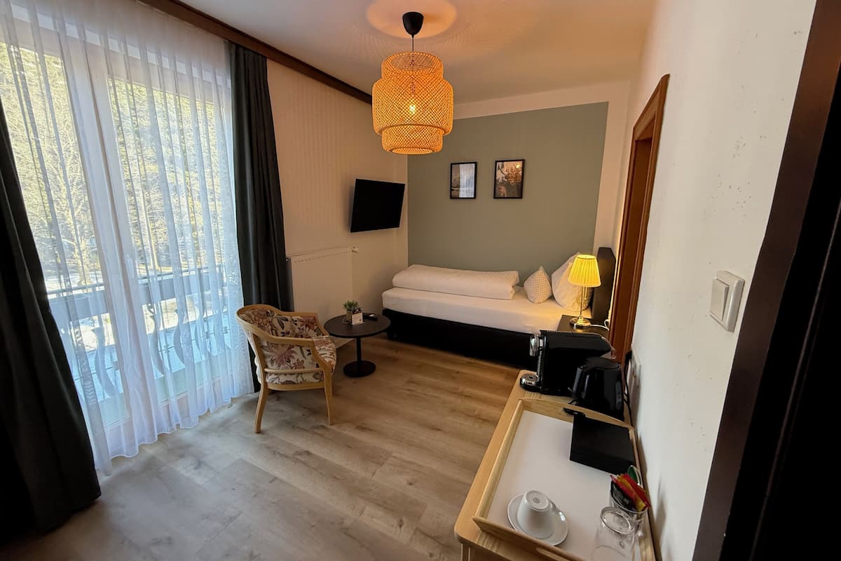 Une chambre d’hôtel avec un plancher en bois, un canapé-lit, une petite table ronde avec une lampe, une chaise, un téléviseur fixé au mur et une fenêtre avec des rideaux légers.