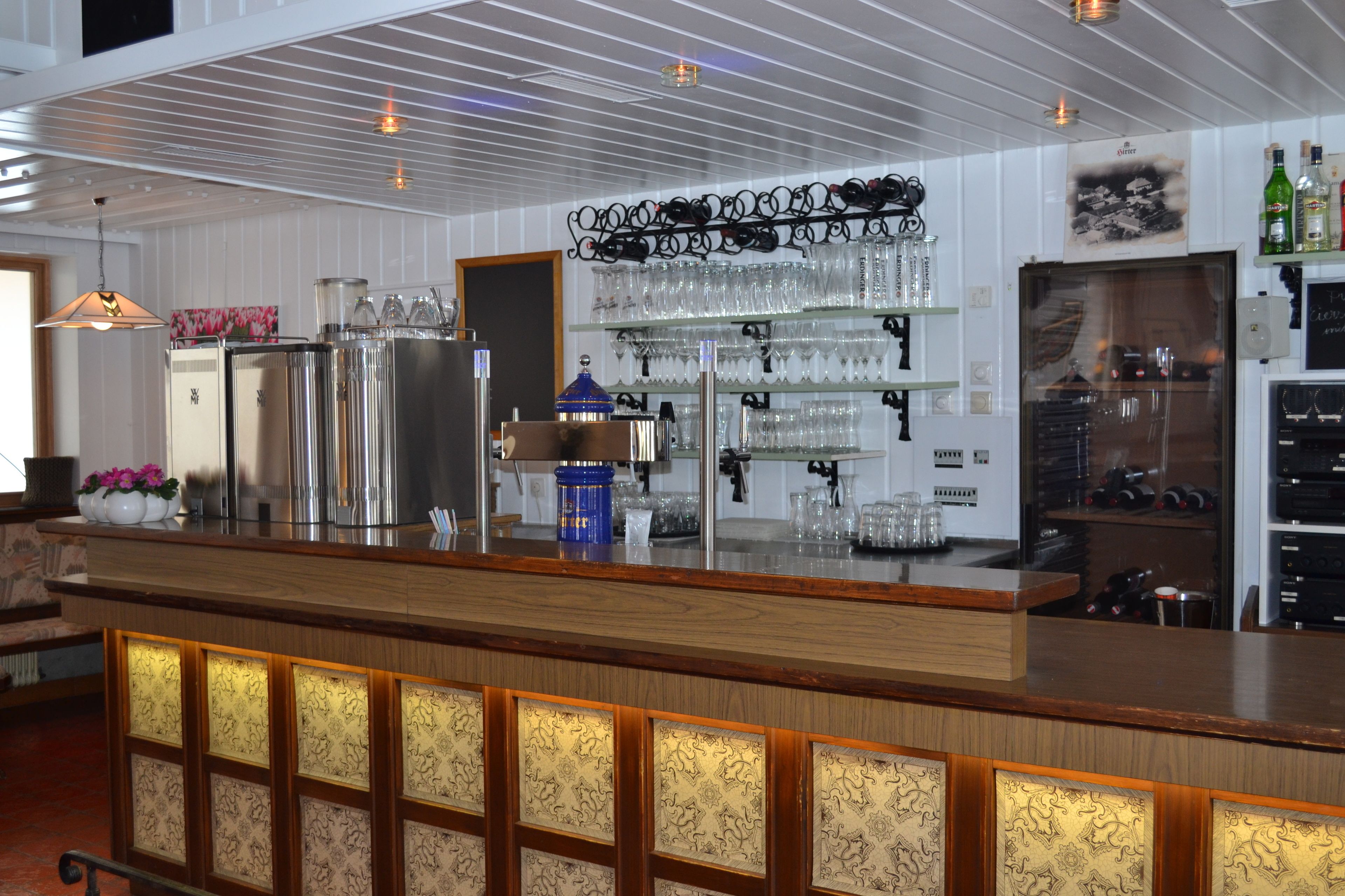 A bar area with a wooden counter, shelves with glasses, a refrigerator, and a blue container.