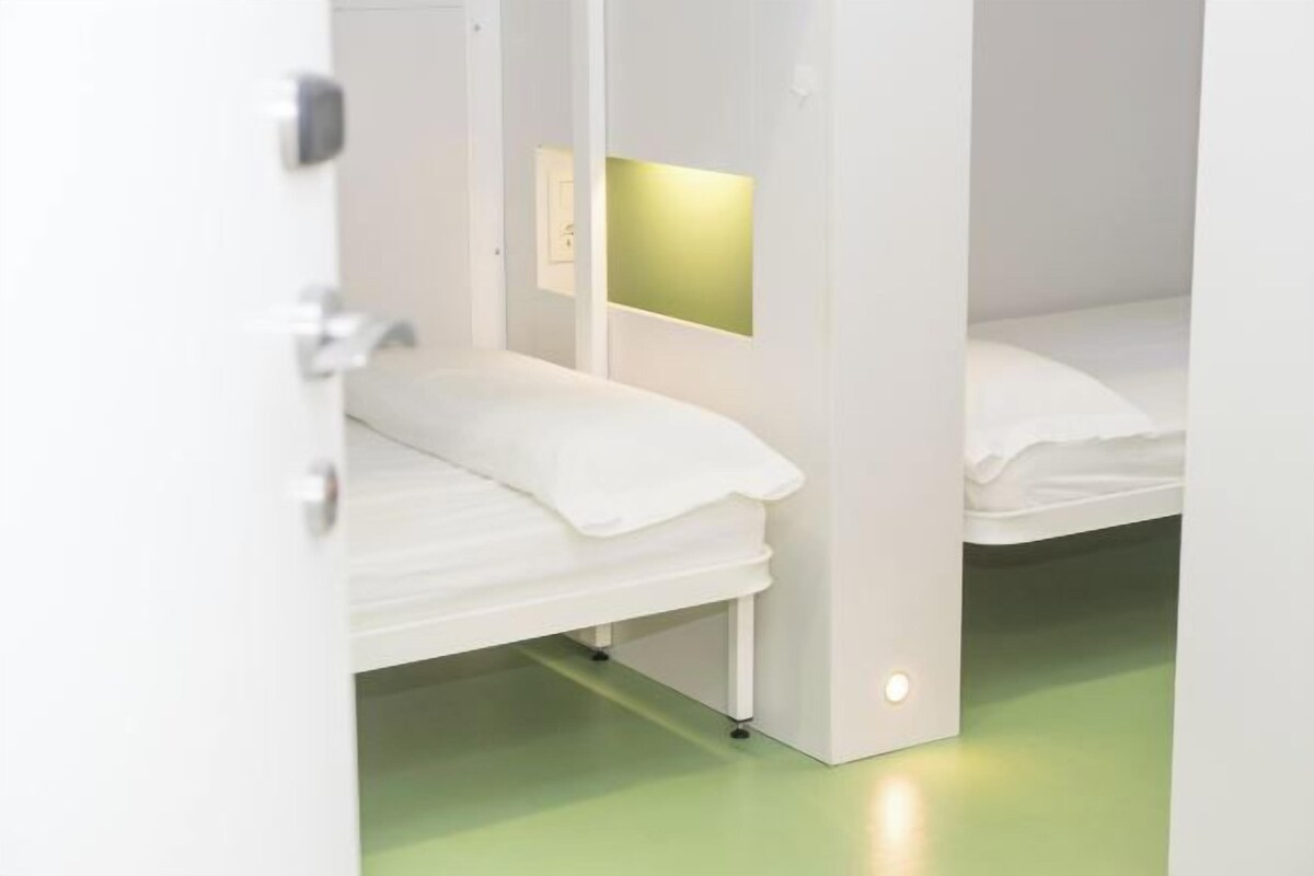 Una habitación de hotel moderna con una cama individual, una mesita de noche y una ventana pequeña con un panel iluminado en verde.