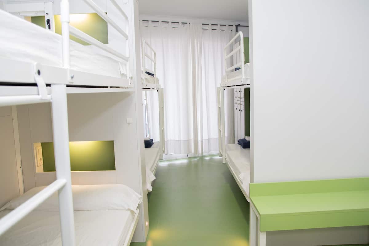A narrow corridor with bunk beds on either side, a green floor, and white curtains at the end.