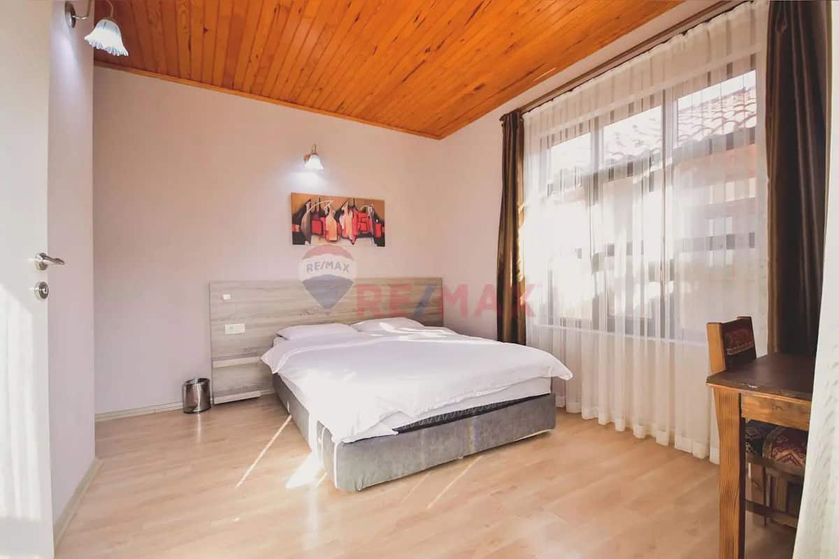A bedroom with a wooden ceiling, a bed with white bedding, a nightstand, a lamp, and a window with sheer curtains.