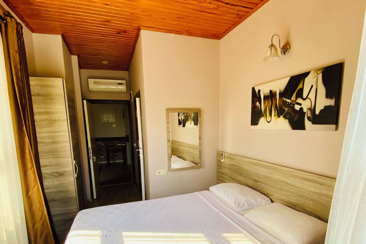 A bedroom with a wooden ceiling, a bed with white linens, a wall-mounted mirror, and a painting.