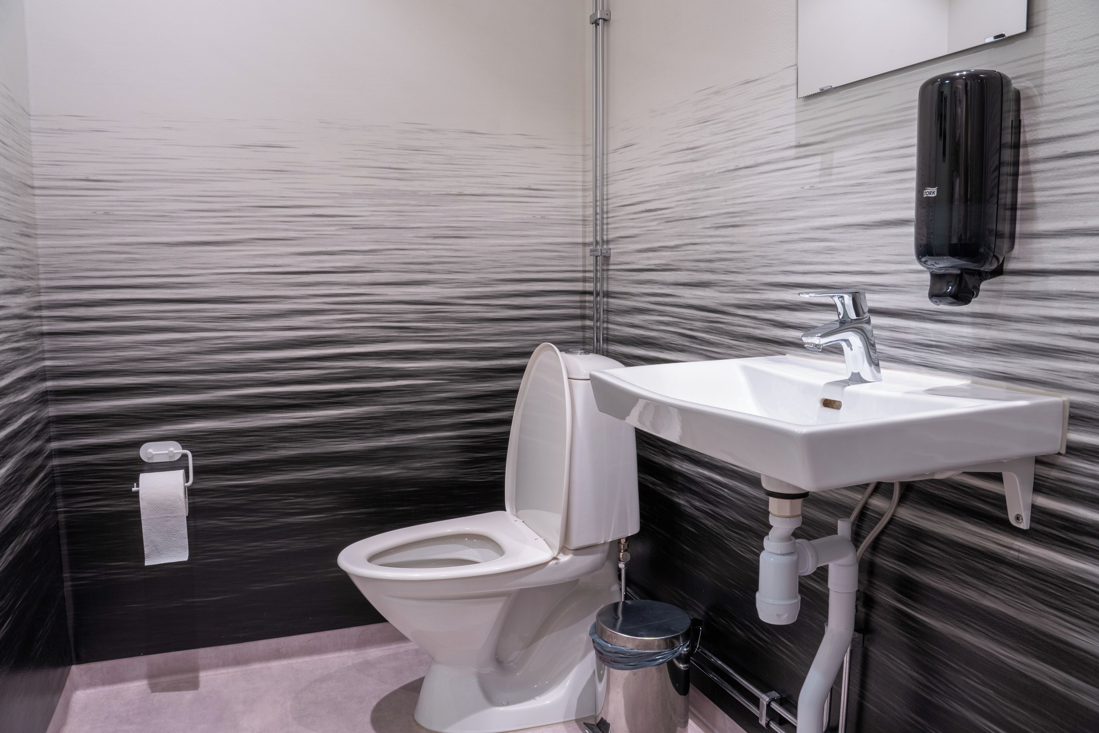 Un baño con inodoro blanco, lavamanos, dispensador de jabón y cubo de basura.