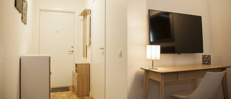 Una habitación compacta con televisión de pantalla plana en la pared, un escritorio pequeño con una silla, un refrigerador y una puerta que da a otra habitación.