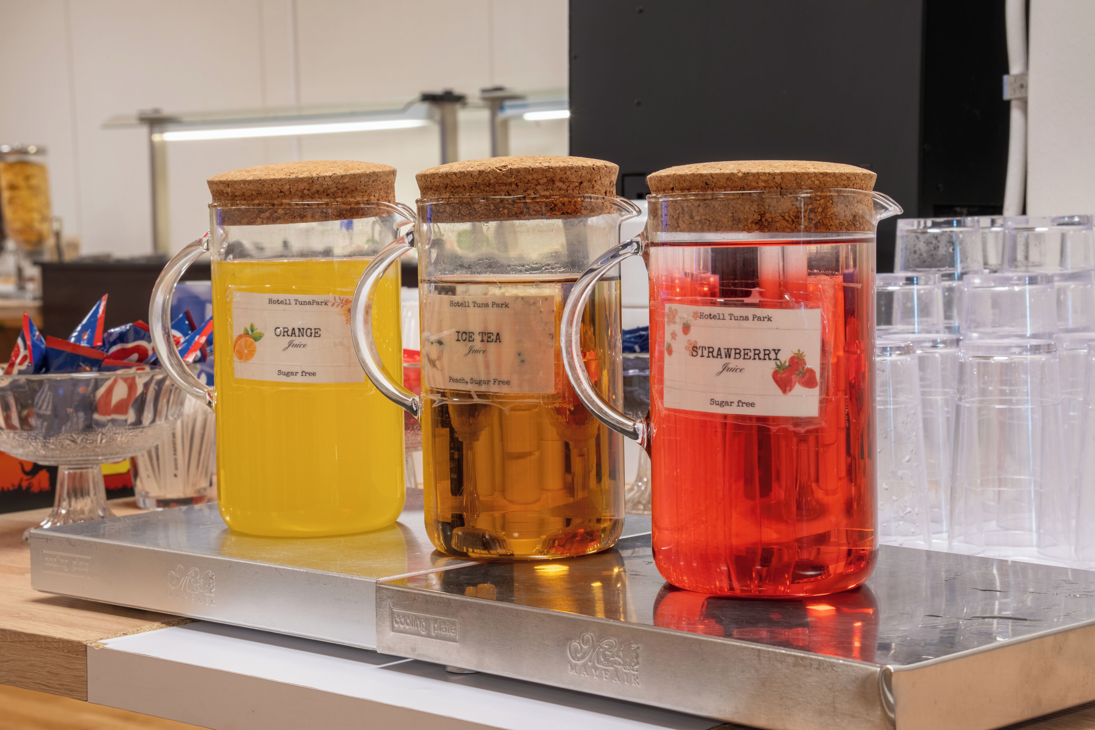Tre glasbehållare med korklock, placerade på en metallbänk, och försedda med etiketter som anger vilken sorts juice de innehåller.