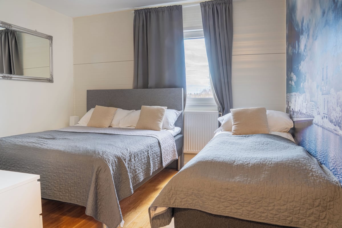 Una cama doble con cabecera gris, dos mesitas de noche, una ventana con cortinas y un espejo fijado a la pared.