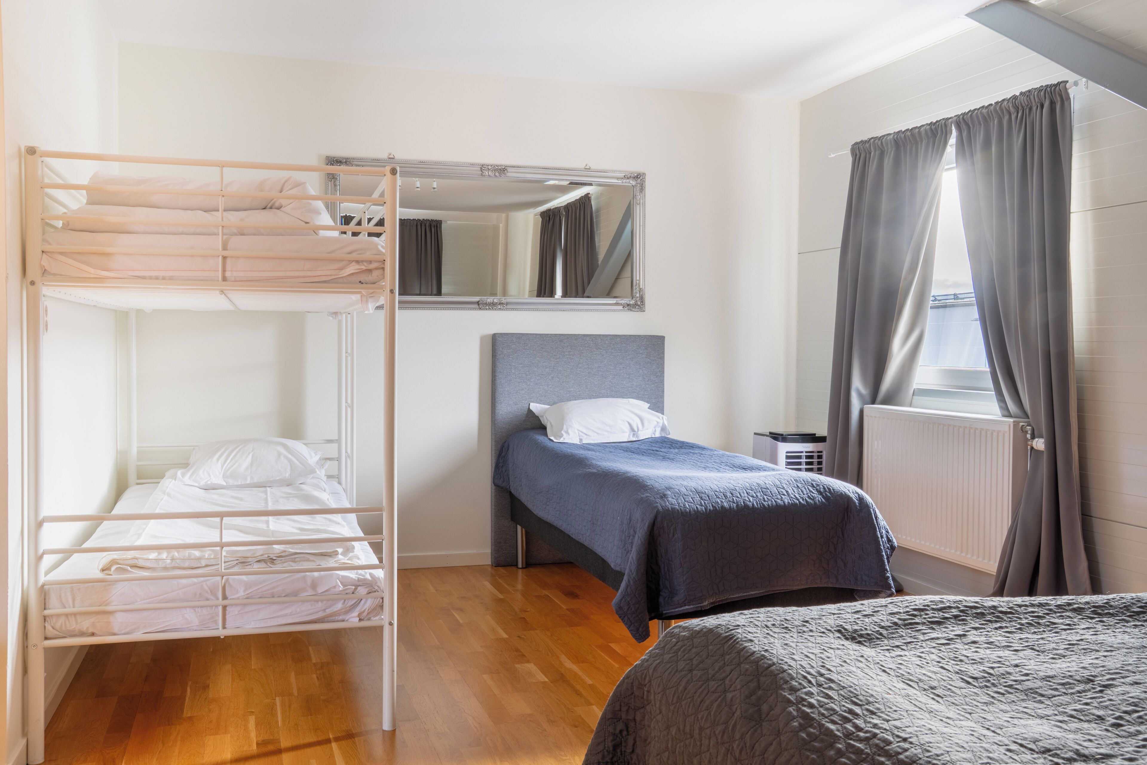 A room with a bunk bed, a single bed, a mirror, a radiator, and a window with curtains.