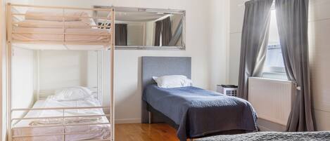 A room with a bunk bed, a single bed, a mirror, a radiator, and a window with curtains.