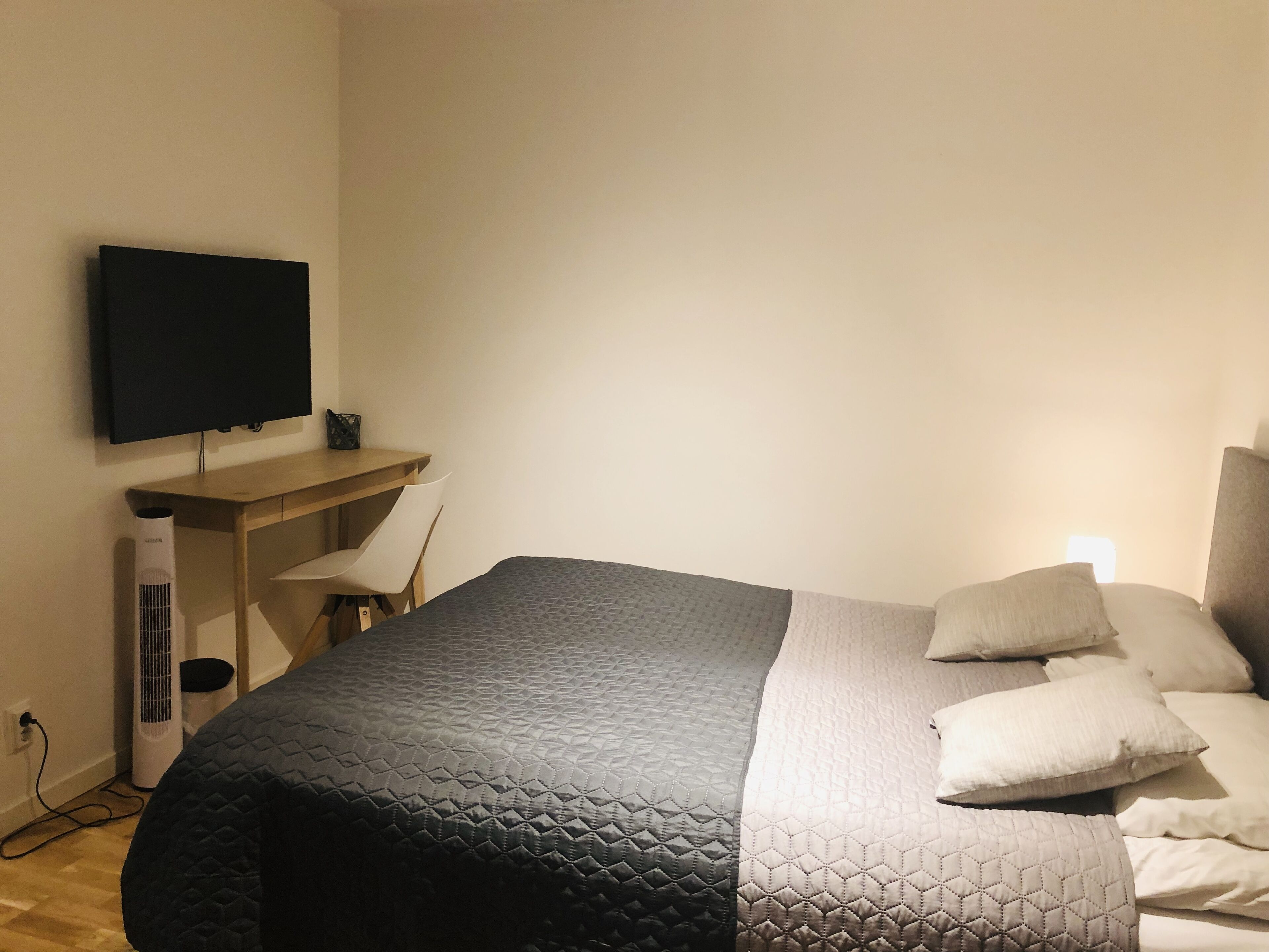 Un dormitorio con cama, almohadas, escritorio, silla y televisor en la pared.