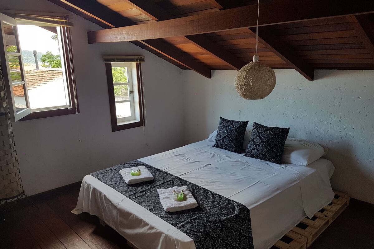 A bedroom with a wooden ceiling, a bed with white bedding, two patterned pillows, and a small window.