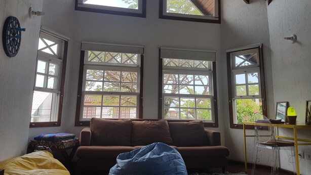 A room with a brown sofa, a blue bean bag, and a desk with a chair. There are three large windows with a view of trees and buildings. - Solar das Pétalas (Florianópolis)