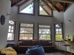 A room with a brown sofa, a blue bean bag, and a desk with a chair. There are three large windows with a view of trees and buildings. - Solar das Pétalas (Florianópolis)