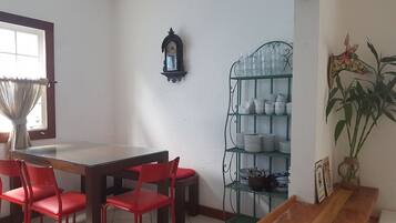Uma área de refeições com mesa de madeira e cadeiras vermelhas, um relógio fixado na parede, uma prateleira de vidro com louças e um vaso de planta.