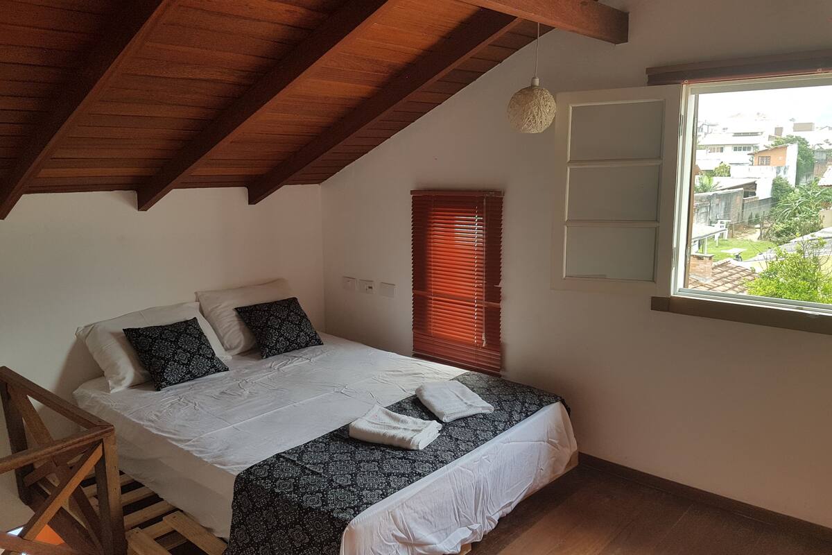 A bedroom with a wooden ceiling, a bed with patterned bedding, a window with blinds, and a view of the outside.