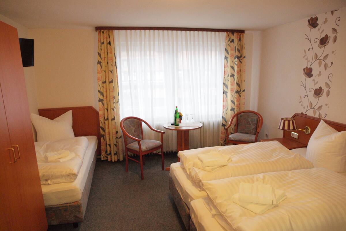 Habitación de hotel con dos camas, una mesita pequeña con una botella y vasos, dos sillas y una mesita de noche con lámpara.