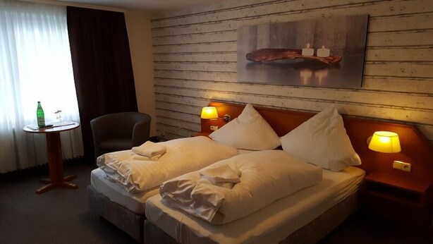 Hotelkamer met een bed, twee bedlampen, een klein tafeltje met een fles, een stoel en een houten wand met een foto.