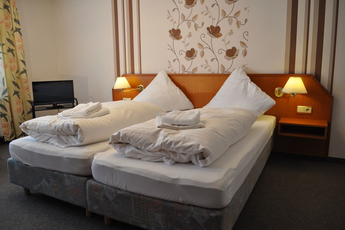 Cama doble con dos almohadas y una toalla doblada, mesita de noche con lámpara, y televisor sobre un mueble.