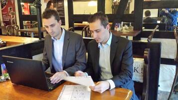 Two men at a table with laptops and documents, in a restaurant with wooden furniture and a menu on the table.