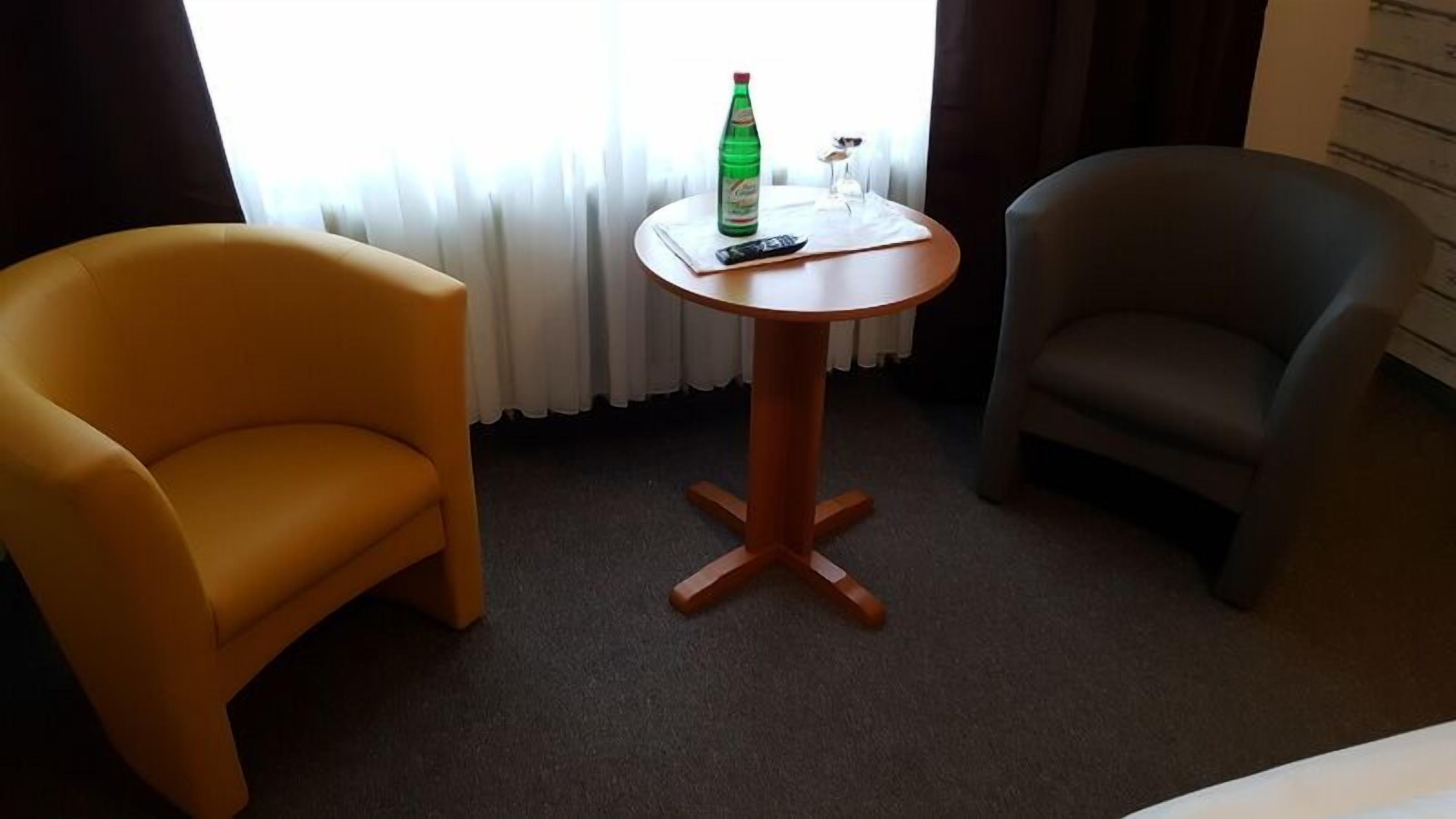 A hotel room with a round wooden table, two armchairs, a bottle of water, and a remote control.