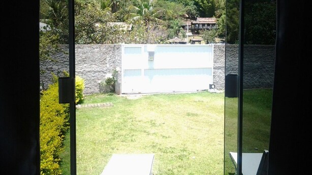 A view from a window overlooking a grassy backyard with a stone wall and a building with multiple windows. - Casa Nunes (Florianópolis)