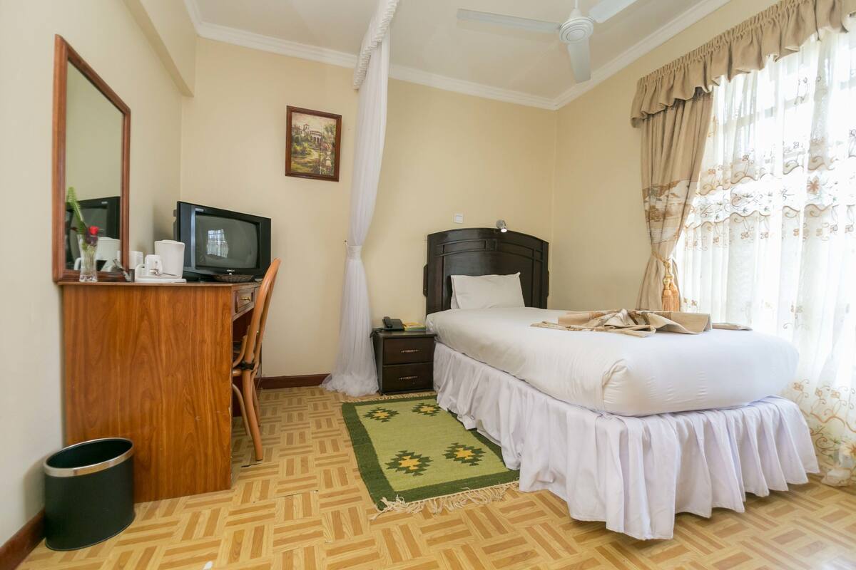 Une chambre d’hôtel comprenant un lit, un bureau avec un ordinateur, une chaise, une petite poubelle et un tapis décoratif.