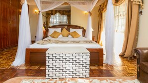 Una cama grande con cabecera de madera, ropa de cama blanca y almohadas doradas. Hay un dosel blanco sobre la cama, y la habitación tiene piso de madera y cortinas.