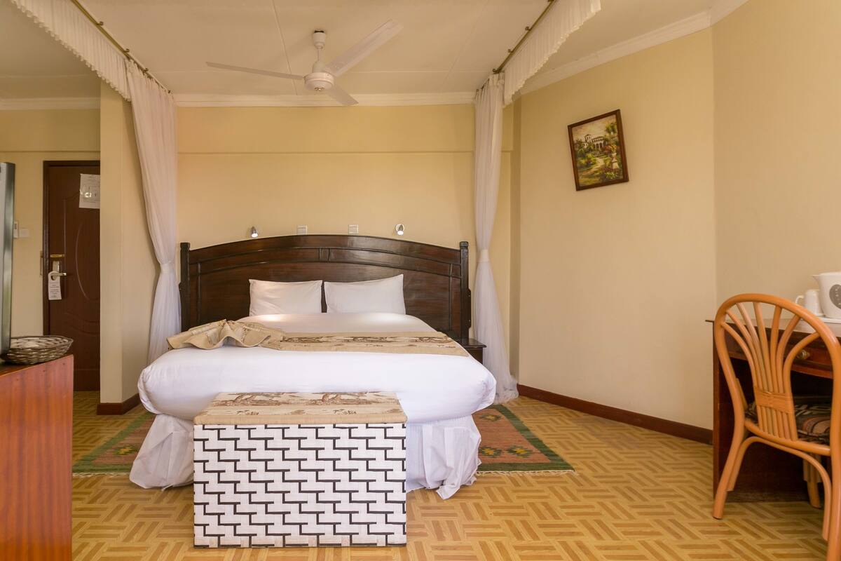 Une chambre à coucher bien rangée, avec un lit en bois, une petite chaise en bois, un ventilateur de plafond et un tableau encadré accroché au mur.