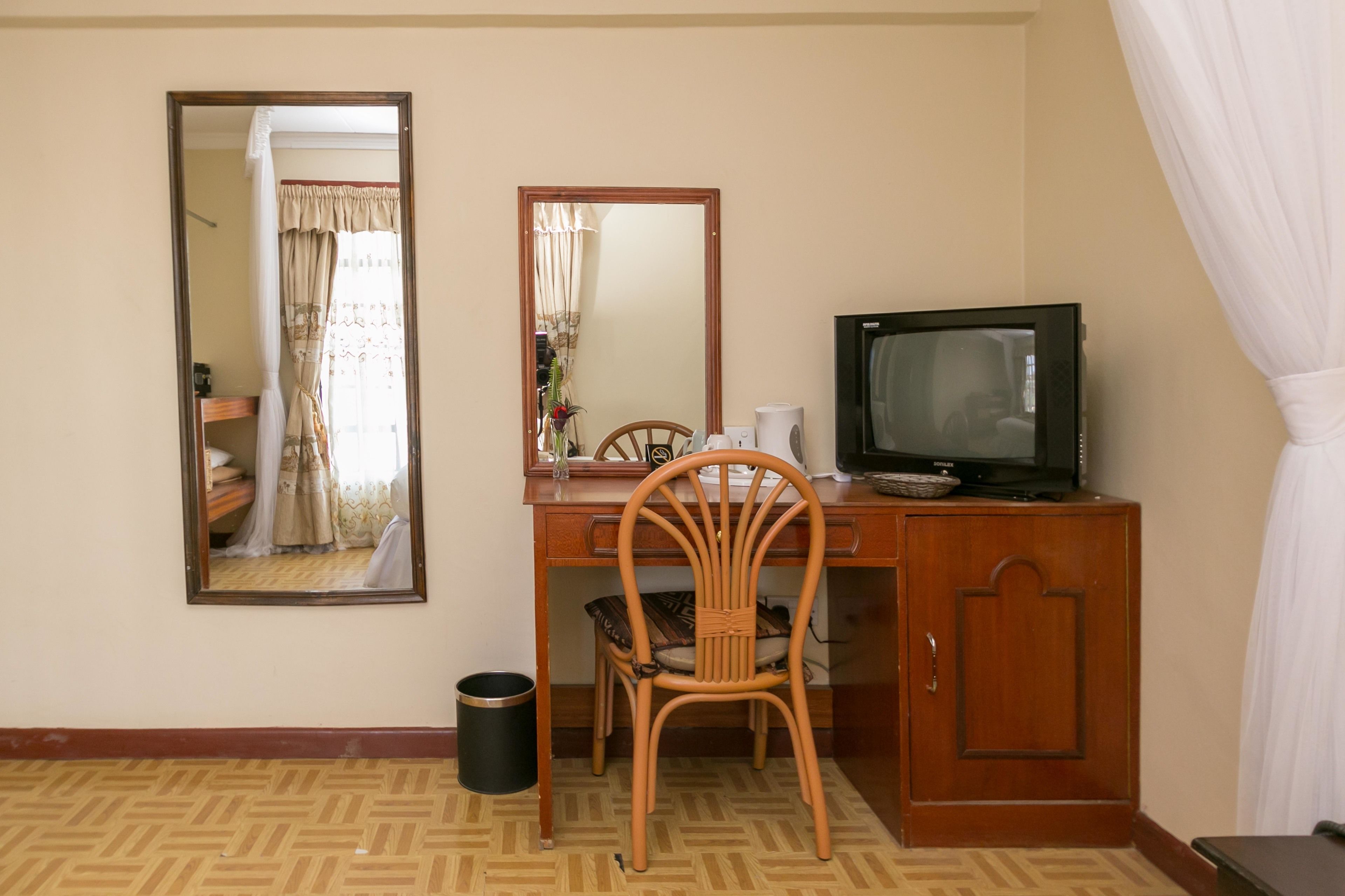A hotel room with a wooden desk, a television, a chair, a mirror, and a waste bin.
