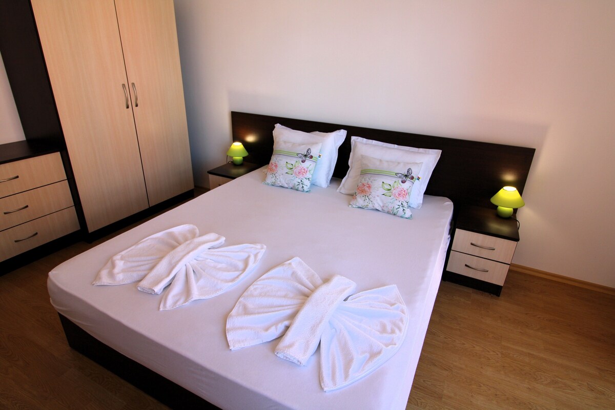 Una cama bien hecha con toallas dobladas en forma de lazo, dos almohadones decorativos con motivos florales y lámparas de noche sobre mesitas de madera.