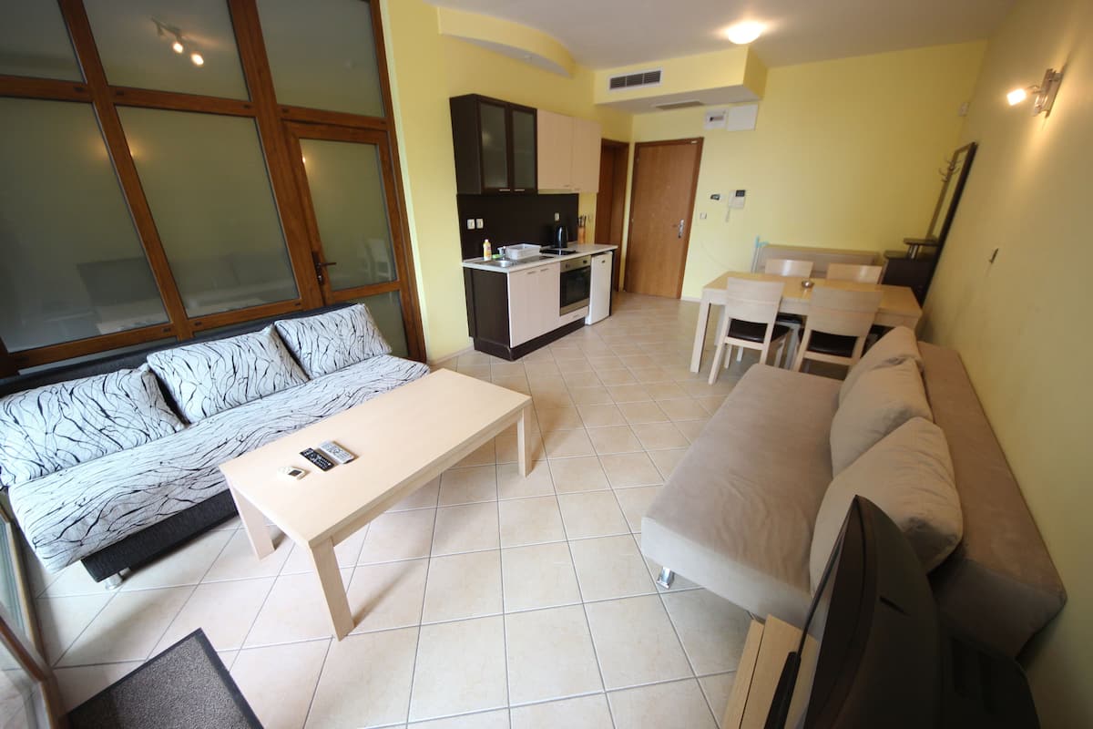A living room with a sofa, a coffee table, a dining table with chairs, a kitchen area with cabinets, and a door.