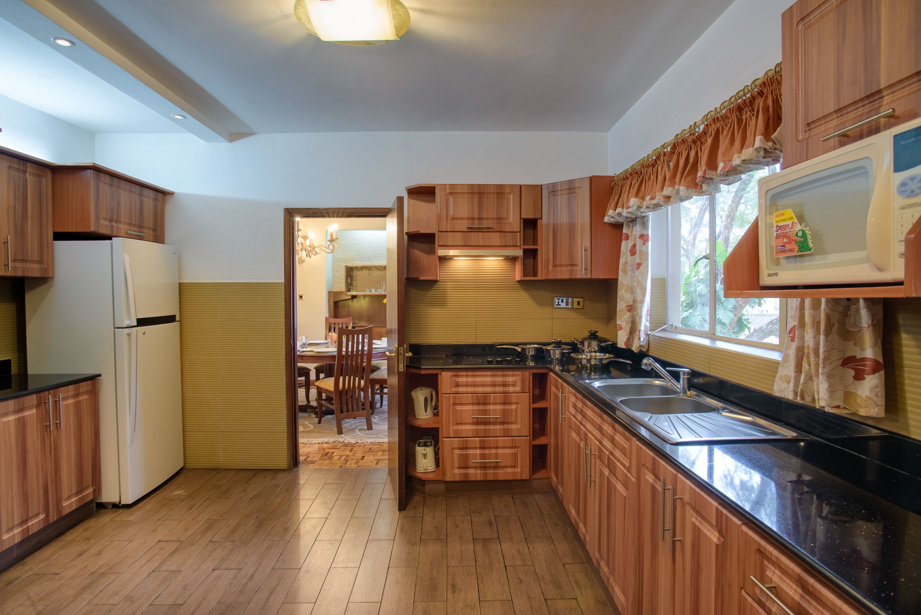 A kitchen with wooden cabinets, a refrigerator, a microwave, and a window with curtains.