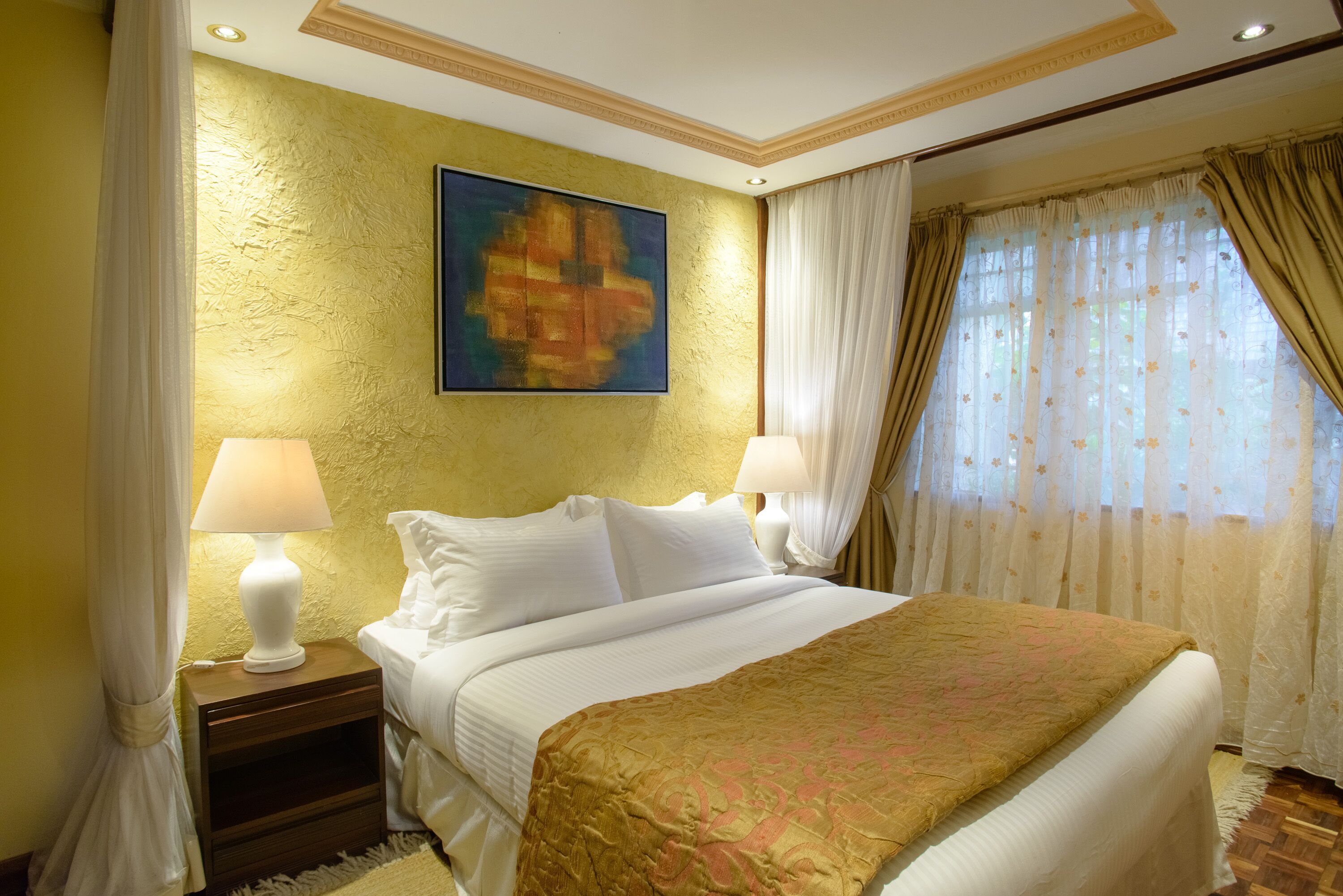 A hotel room with a large bed, two bedside lamps, a gold textured wall, and a painting above the bed.