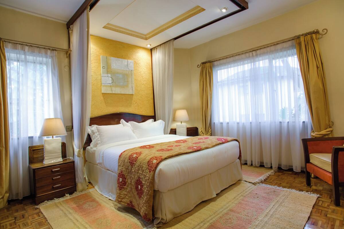 A hotel room with a large bed, two bedside tables with lamps, a wooden headboard, and a view of the outdoors through large windows with curtains.