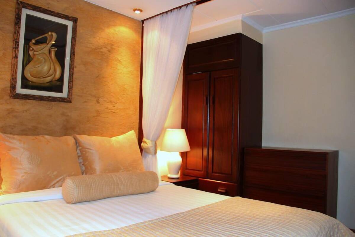 A hotel room with a bed, pillows, a nightstand, a lamp, a wooden wardrobe, and a framed picture on the wall.