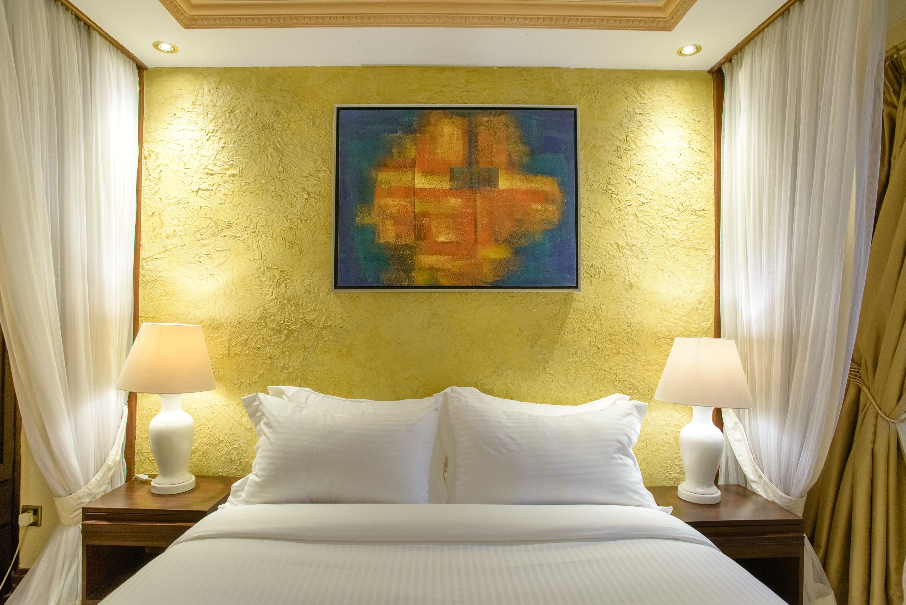 A hotel room with a bed, two bedside tables with lamps, white bedding, and a textured yellow wall.