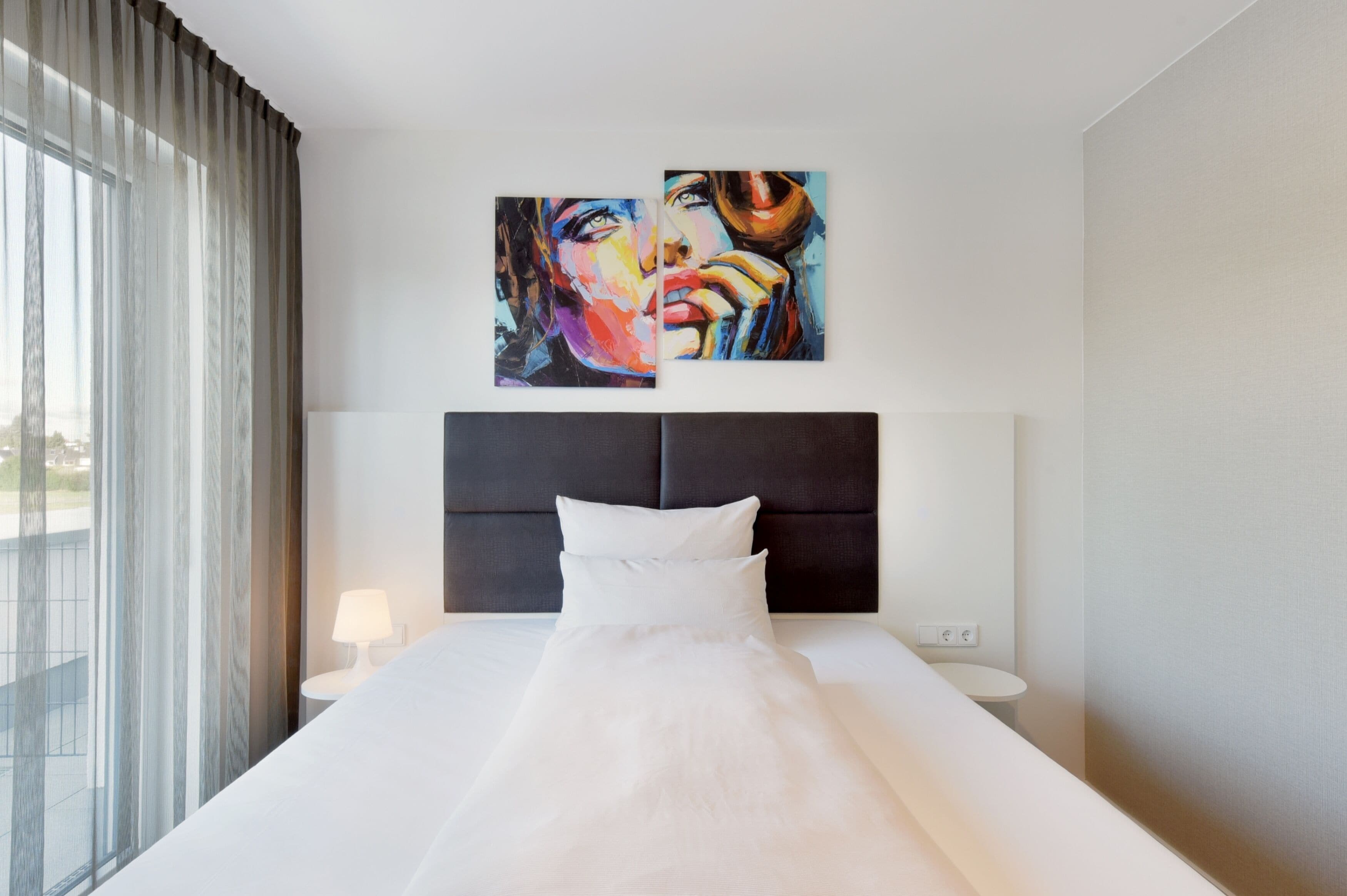 A hotel room with a large bed, a headboard, a bedside table with a lamp, and two paintings on the wall.