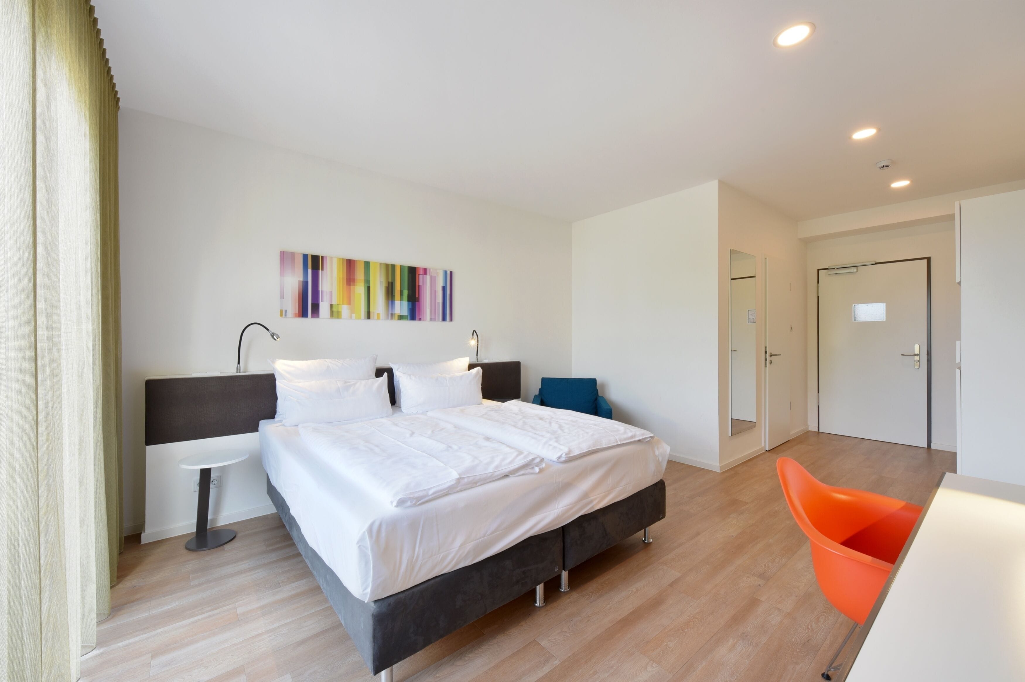 Habitació d'hotel moderna amb un llit gran, una petita taula de nit, una cadira i una porta que condueix a una altra habitació.
