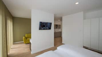 A hotel room with a bed, a TV mounted on the wall, a yellow chair, and a kitchenette in the background.