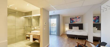 A modern hotel room with a glass shower, a wooden vanity, a bed with a headboard, and a desk with a chair.