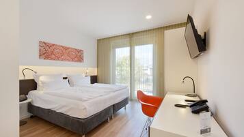 Habitación de hotel con cama, escritorio con silla, televisor en la pared y ventana con cortinas.