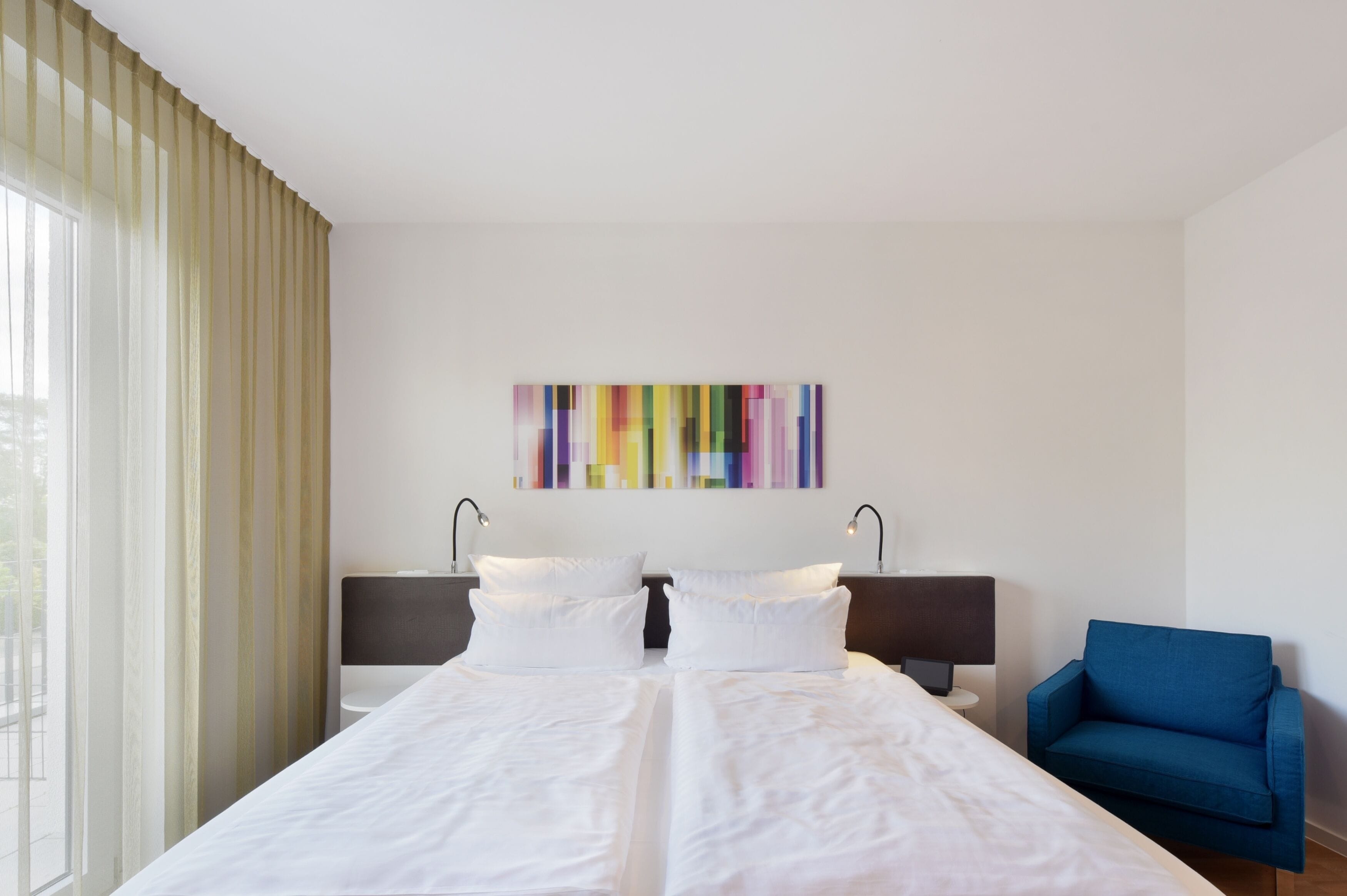 A hotel room with a large bed, a blue armchair, and a colorful abstract painting on the wall.