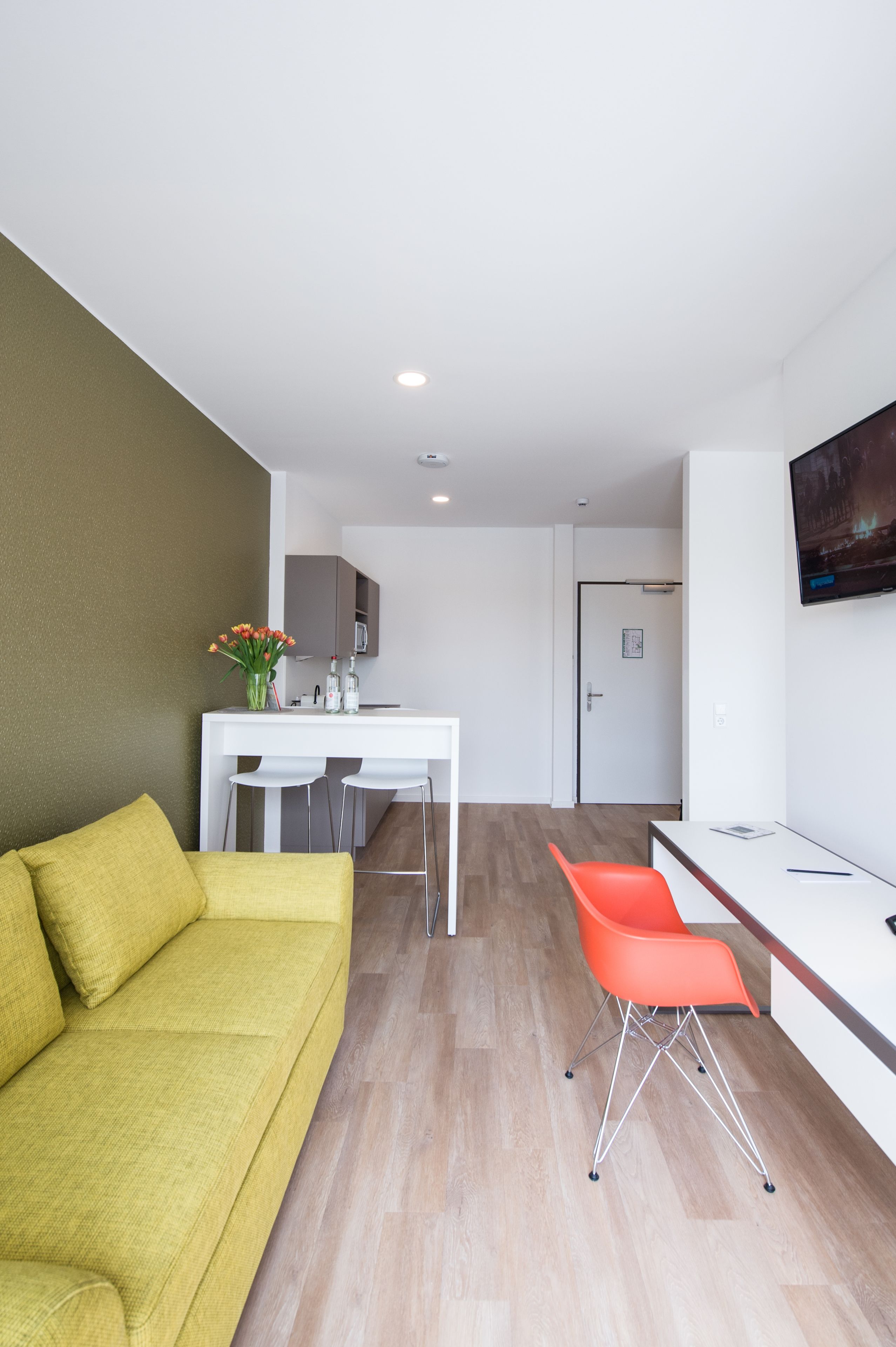 Un salon moderne avec un canapé jaune, un coin repas avec une table et des chaises blanches, et une cuisine en arrière-plan.