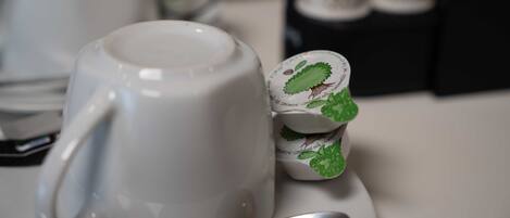 Primer plano de una taza blanca con una cuchara sobre un platito, una pila de tazas pequeñas con diseño de hoja verde, y una caja negra al fondo.