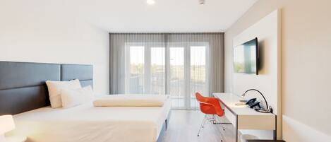 A hotel room with a bed, desk, chair, and a large window.