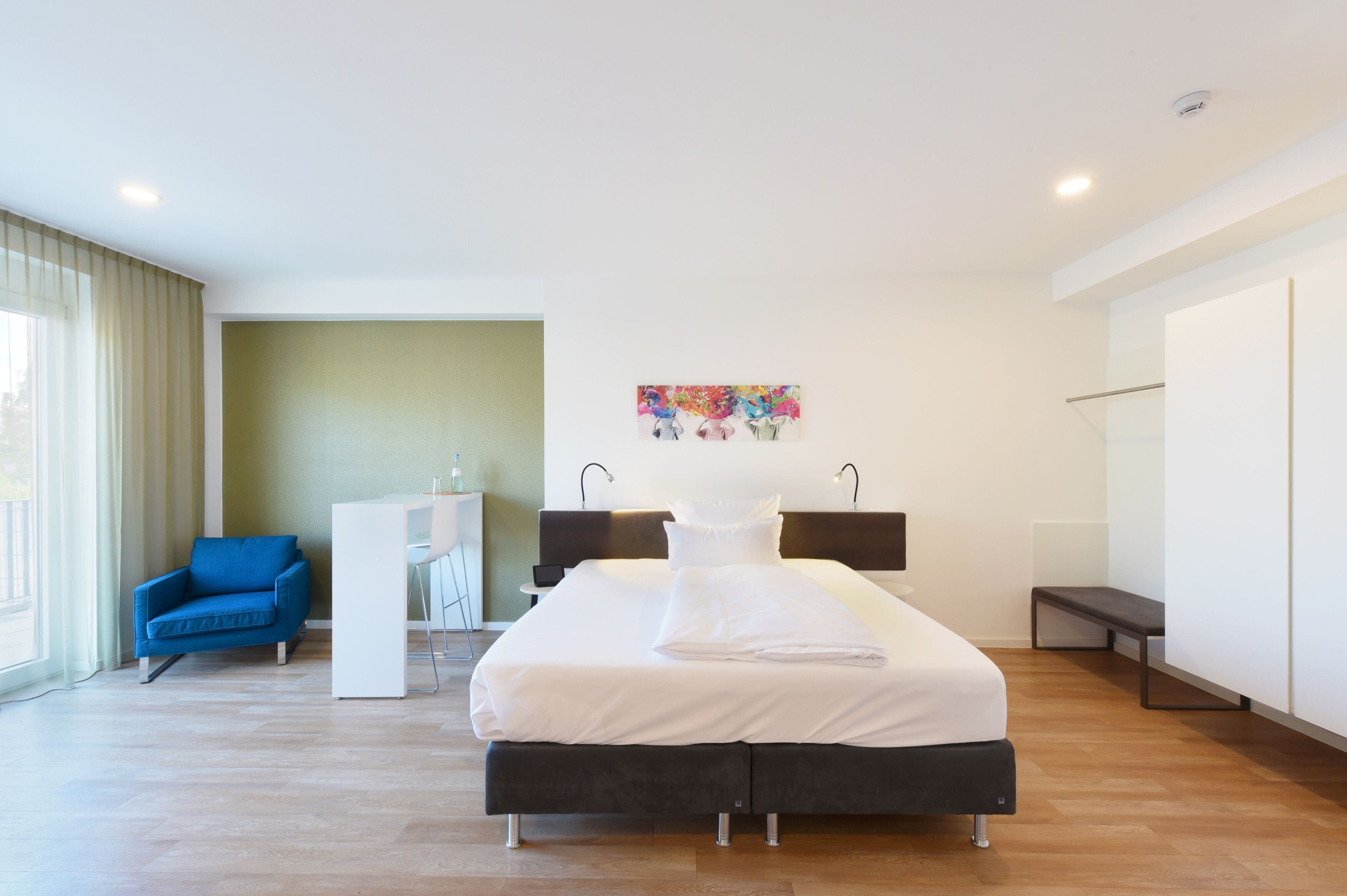 Ein modernes Hotelzimmer mit einem großen Bett, einem blauen Sessel, einem Schreibtisch und einem farbenfrohen Kunstwerk an der Wand.