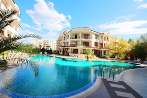 A swimming pool with a tiled edge, surrounded by lounge chairs and palm trees, with a multi-story building in the background. - Menada Chateau Valon Apartments (Sunny Beach)