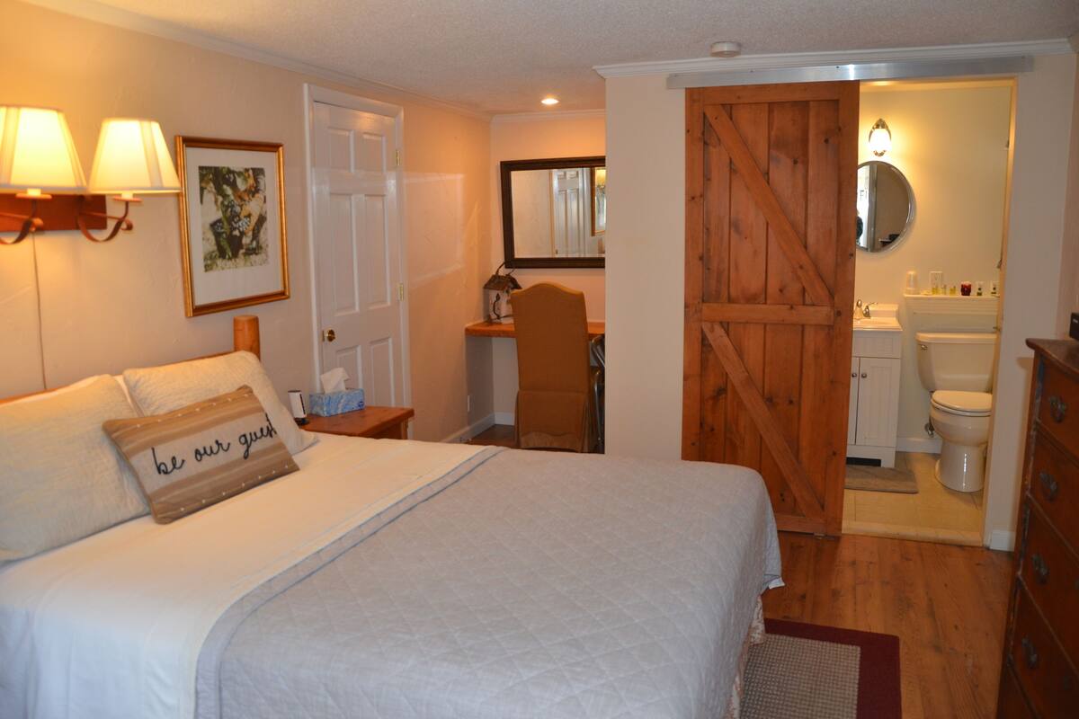 A bedroom with a bed, bedside lamps, a desk, a chair, and a wooden door leading to a bathroom.