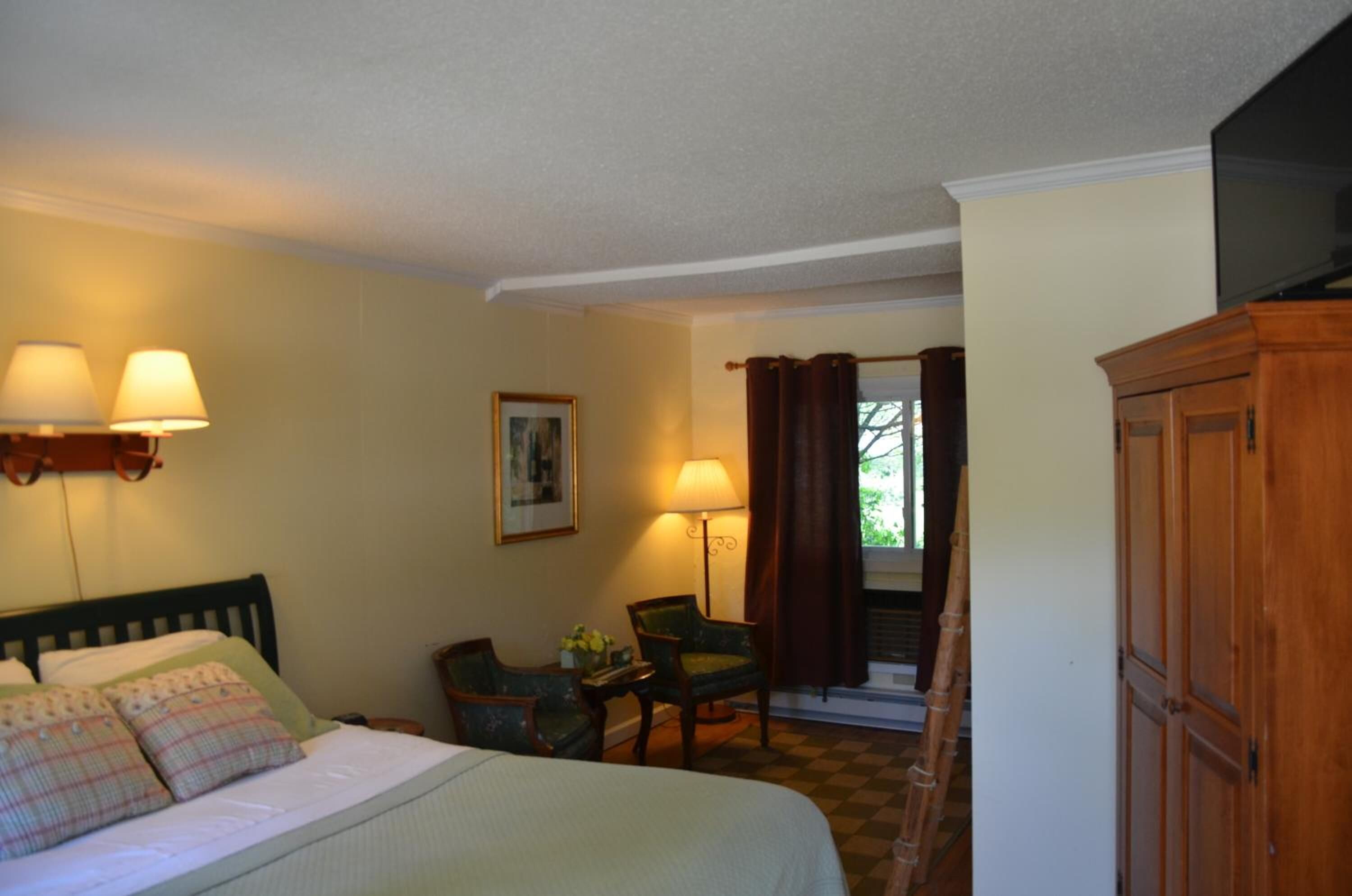 A hotel room with a bed, two chairs, a desk, a lamp, a picture on the wall, and a wardrobe.