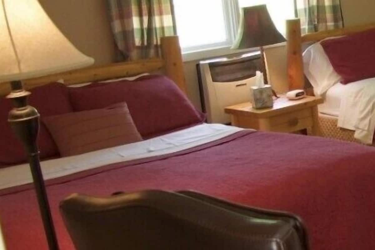 A hotel room with two beds, each with a red bedspread, a bedside table with a lamp, and a window with curtains.